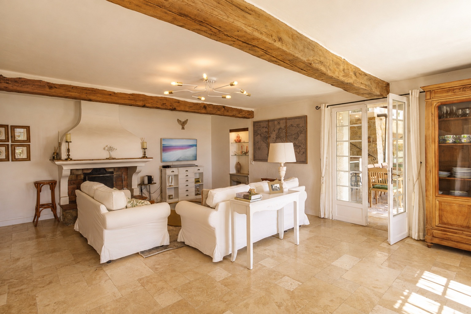 White sofas beneath exposed timber beams, with a fireplace and open doors to the terrace